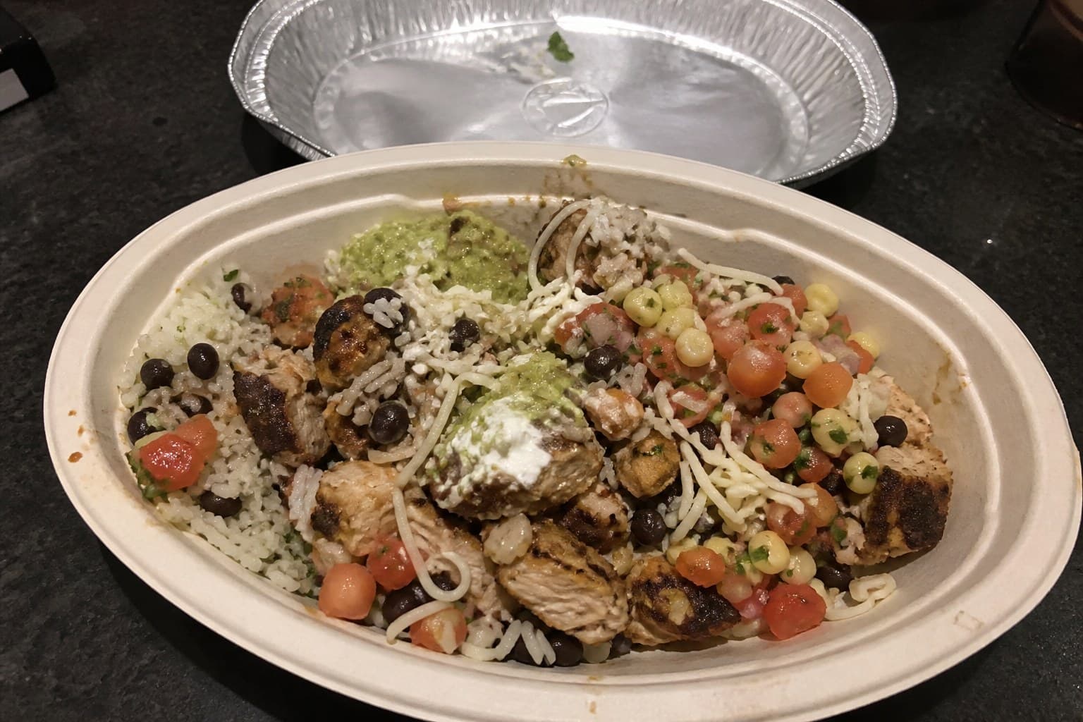 A Chipotle-style burrito bowl in a compostable fiber takeout bowl with rice, chicken, beans, salsa, cheese, and guacamole.