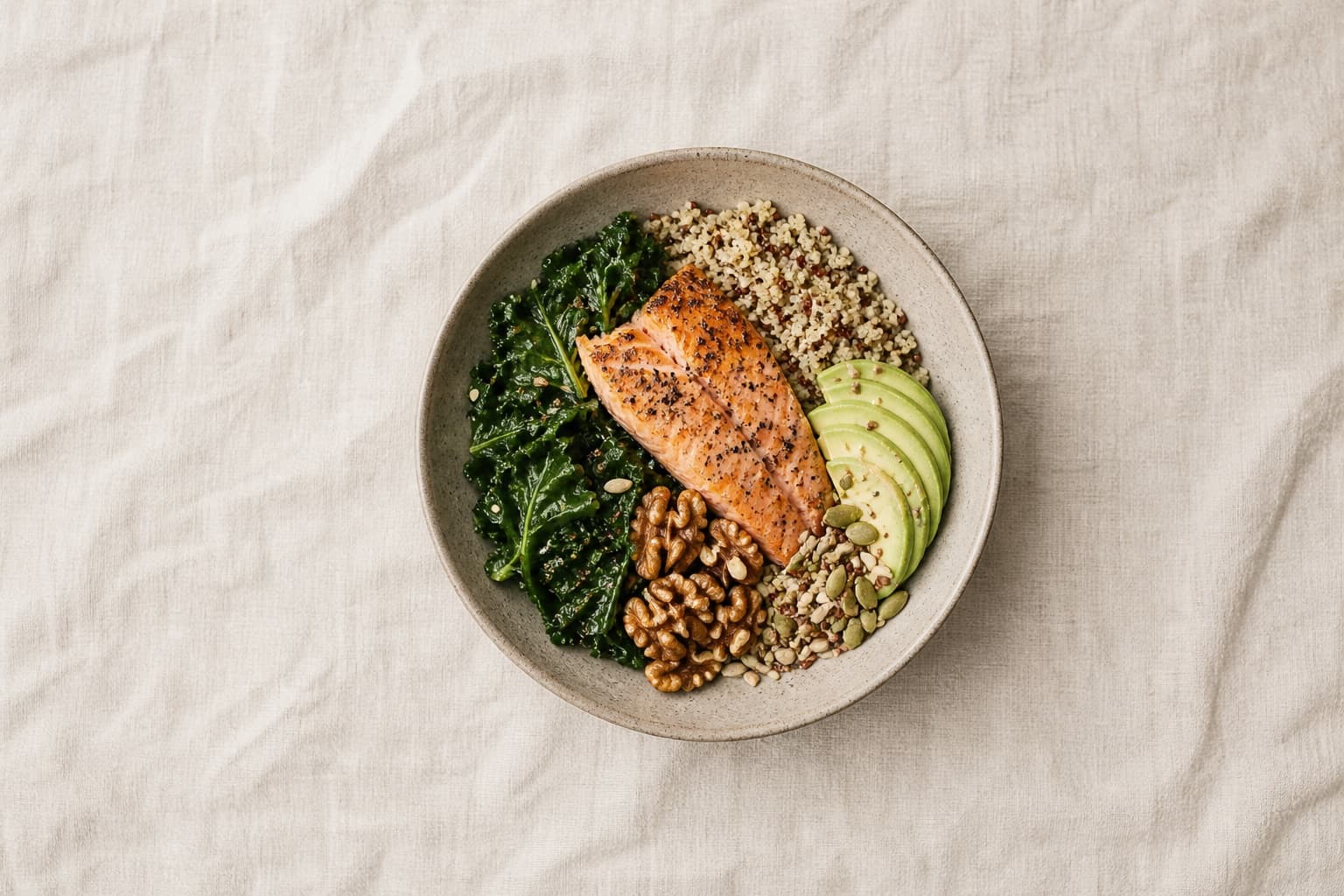 An overhead flatlay of colorful whole foods on a cream linen surface: leafy greens, citrus, nuts, salmon, and eggs arranged in bowls.