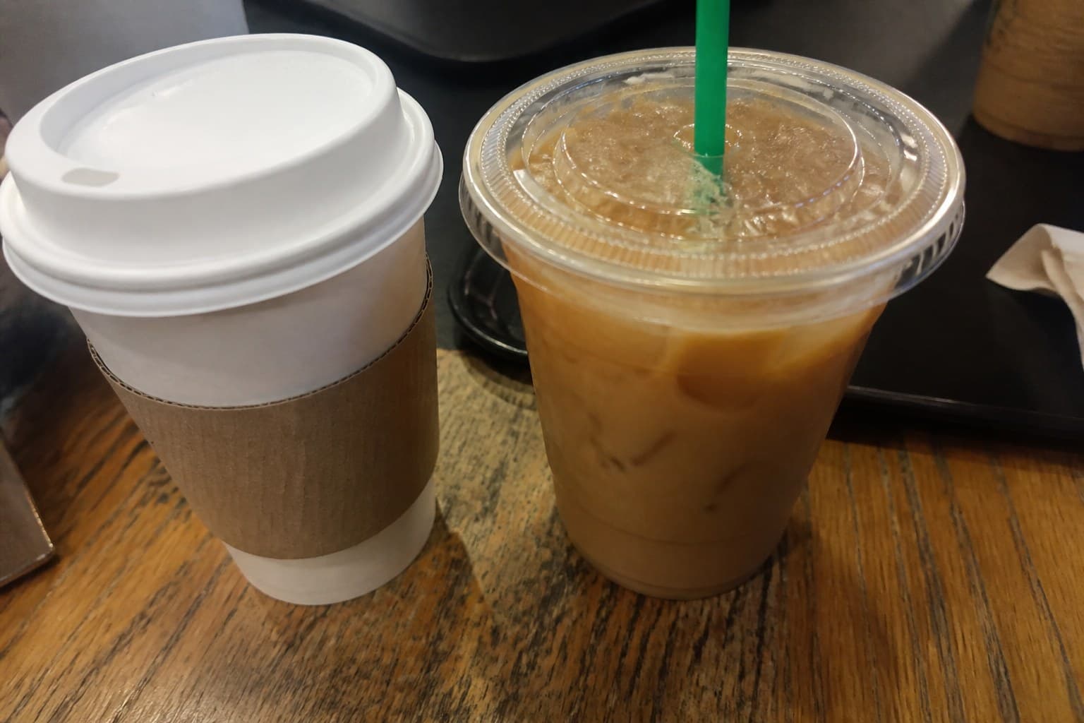 A hot Starbucks-style paper cup beside a clear iced coffee cup with ice and a green straw on a cafe table.